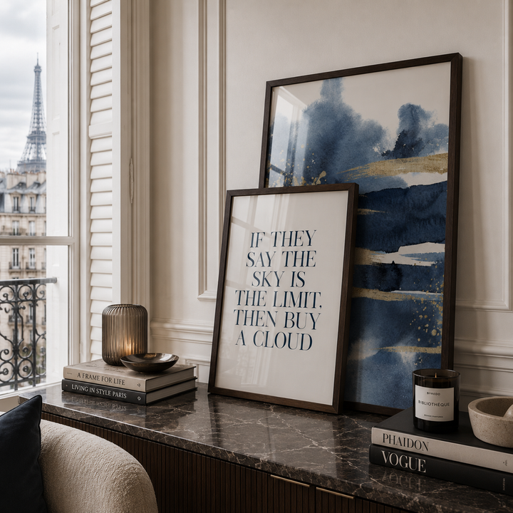 Two framed art prints displayed on a dark marble console table in a styled interior. The front print features a quote reading 'IF THEY SAY THE SKY IS THE LIMIT, THEN BUY A CLOUD' in dark blue text on a white background.