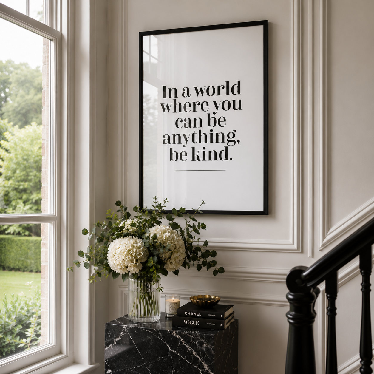 A framed typographic wall art print with the quote 'In a world where you can be anything, be kind.' hangs on a cream paneled wall above a black marble side table with white flowers and books.