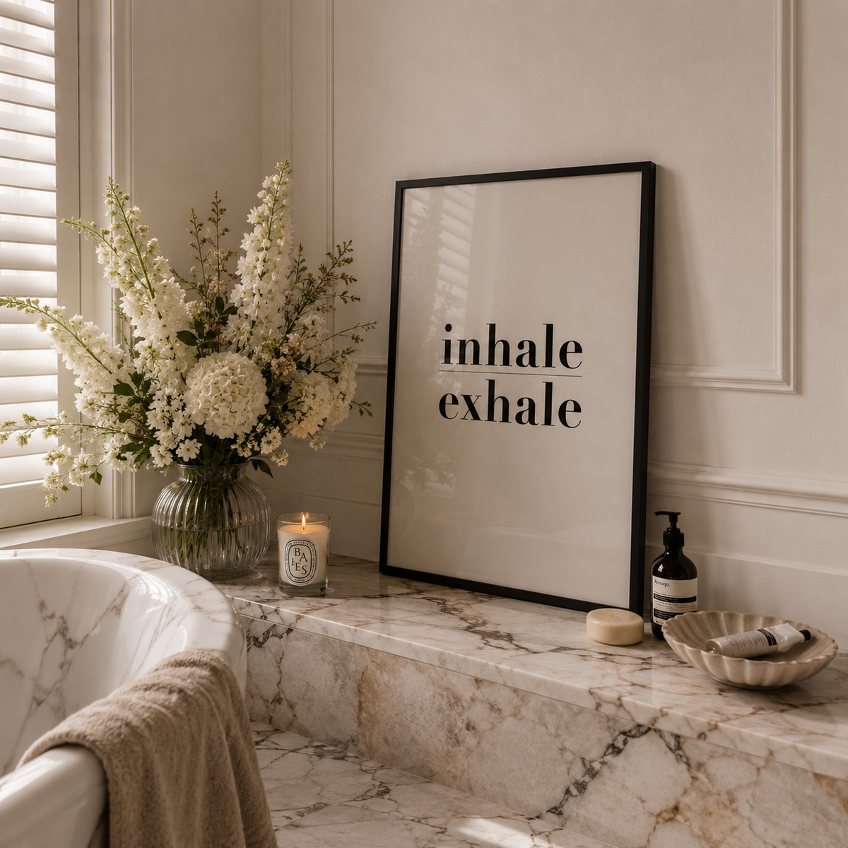 A framed minimalist art print with the words 'inhale exhale' in black serif text on a warm off-white background, leaning on a marble bathroom counter next to a large floral arrangement and a lit candle.