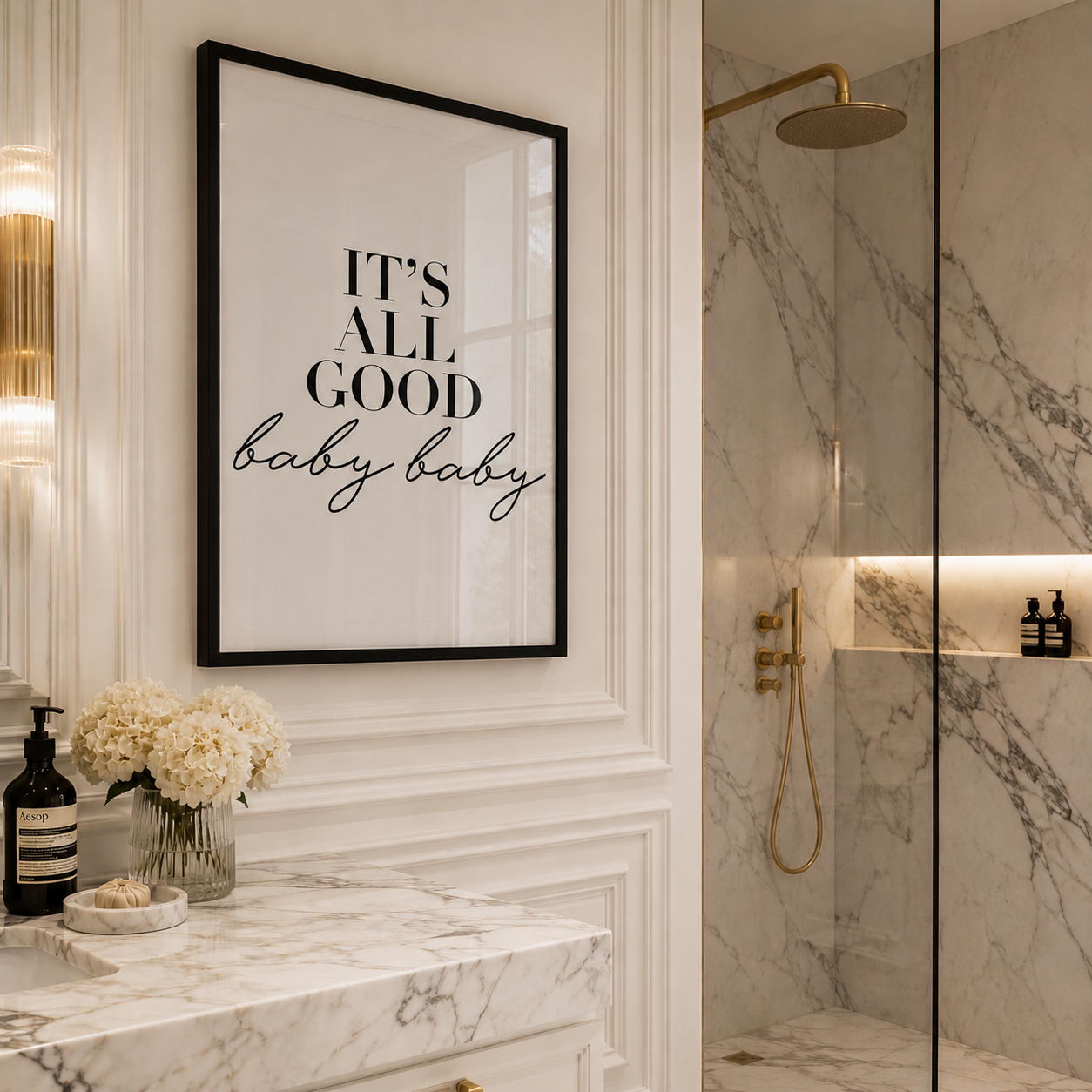 Framed wall art with the text 'IT'S ALL GOOD baby baby' in black lettering on a white background, hanging in a modern bathroom with marble surfaces and gold fixtures.