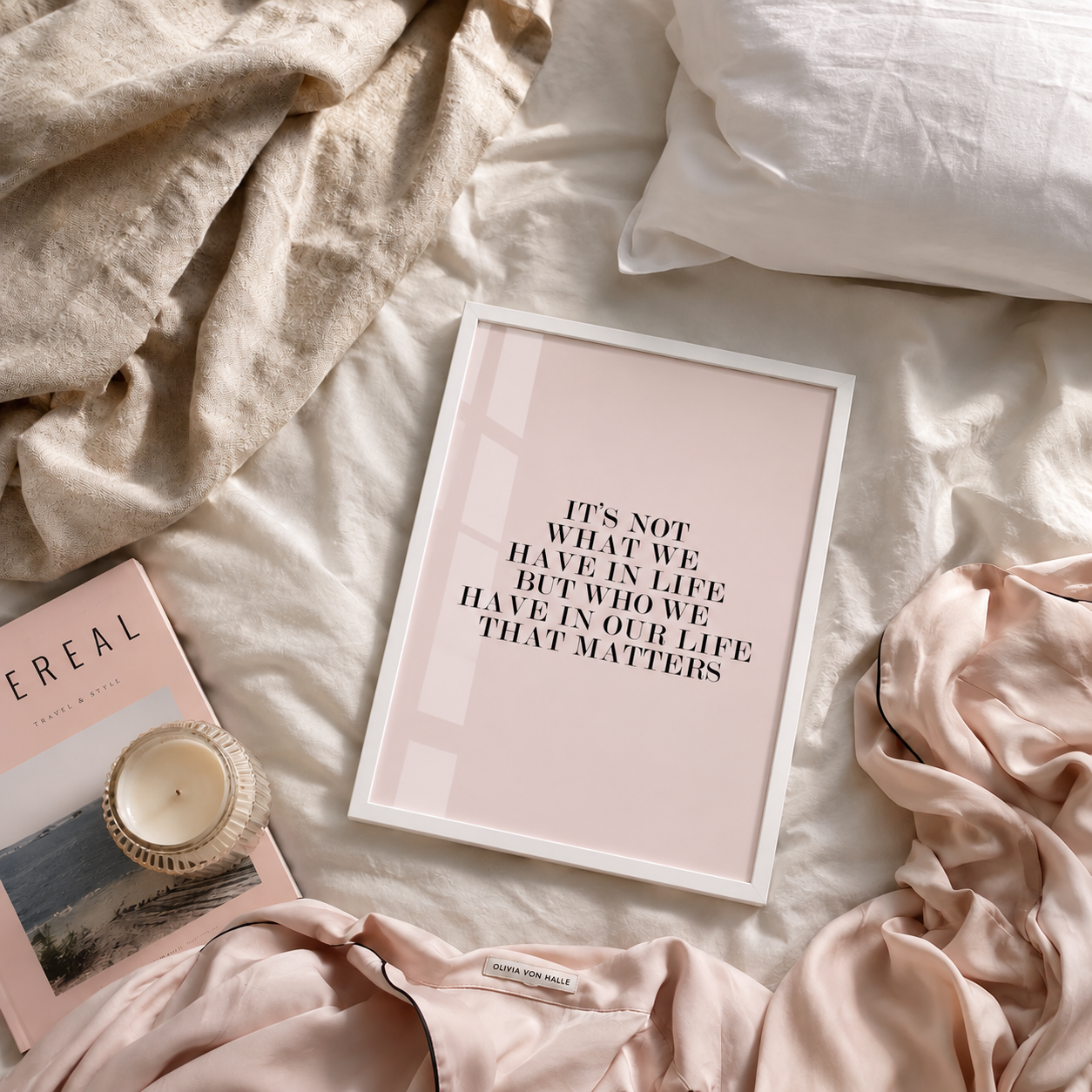 A framed typographic art print with the quote 'IT'S NOT WHAT WE HAVE IN LIFE BUT WHO WE HAVE IN OUR LIFE THAT MATTERS' in black text on a pale blush pink background, styled on a bed with beige linens, a pink cereal box, a candle, and soft blush fabric.