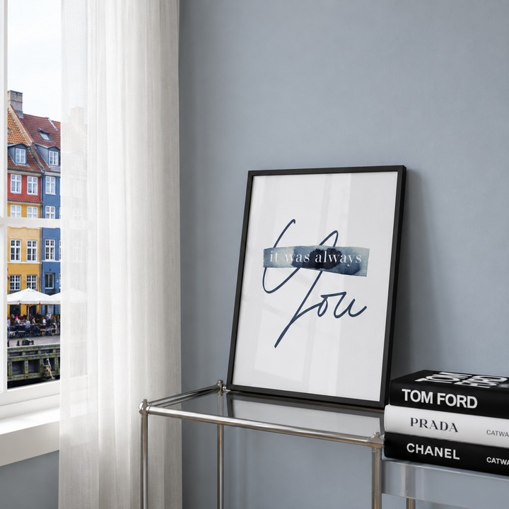 A framed wall art print with the phrase 'It was always You' in dark blue script and a textured blue-gray banner, leaning on a glass side table in a room with a gray wall and a window view of colorful buildings.