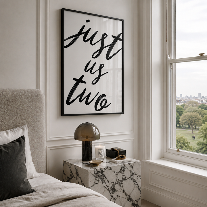 A framed wall art print with the phrase 'just us two' in black script on a white background, hanging above a marble nightstand in a styled bedroom.