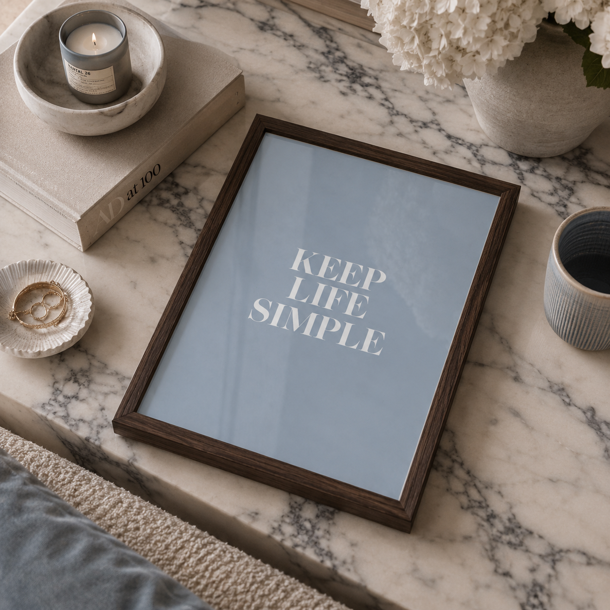 A framed art print with the text 'KEEP LIFE SIMPLE' in white on a light blue background, styled on a white and gray marble surface with books, a candle, flowers, and a cup.