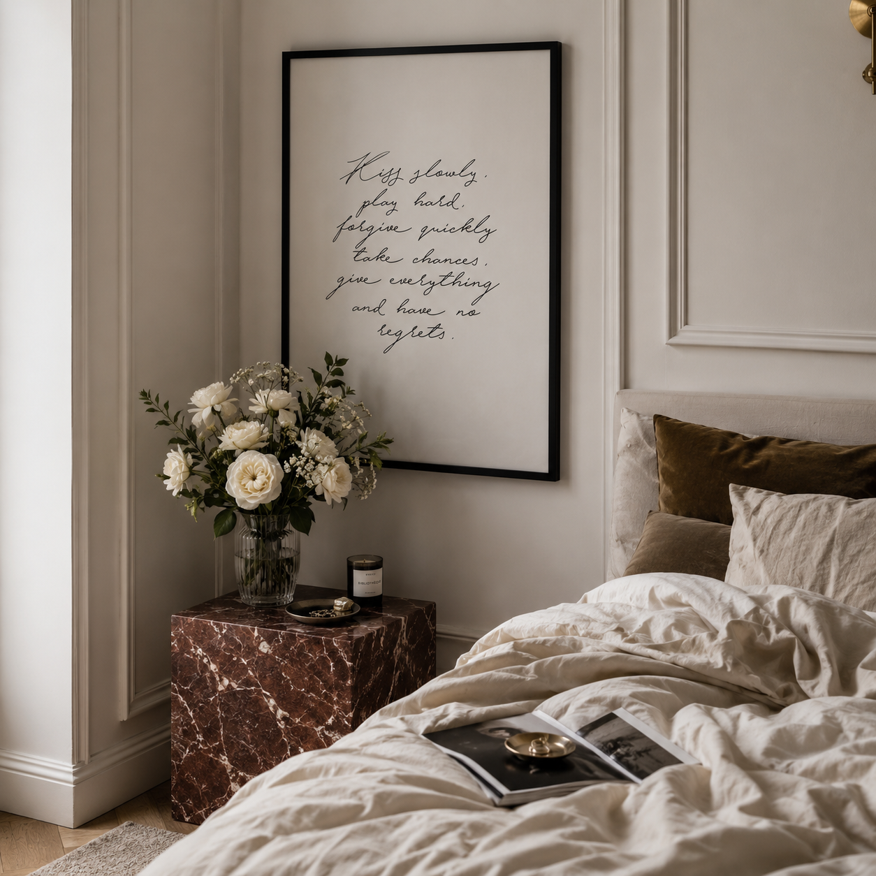 A framed wall art print with elegant cursive script hanging above a bed in a styled bedroom. The print has a black frame and a white background with handwritten text.