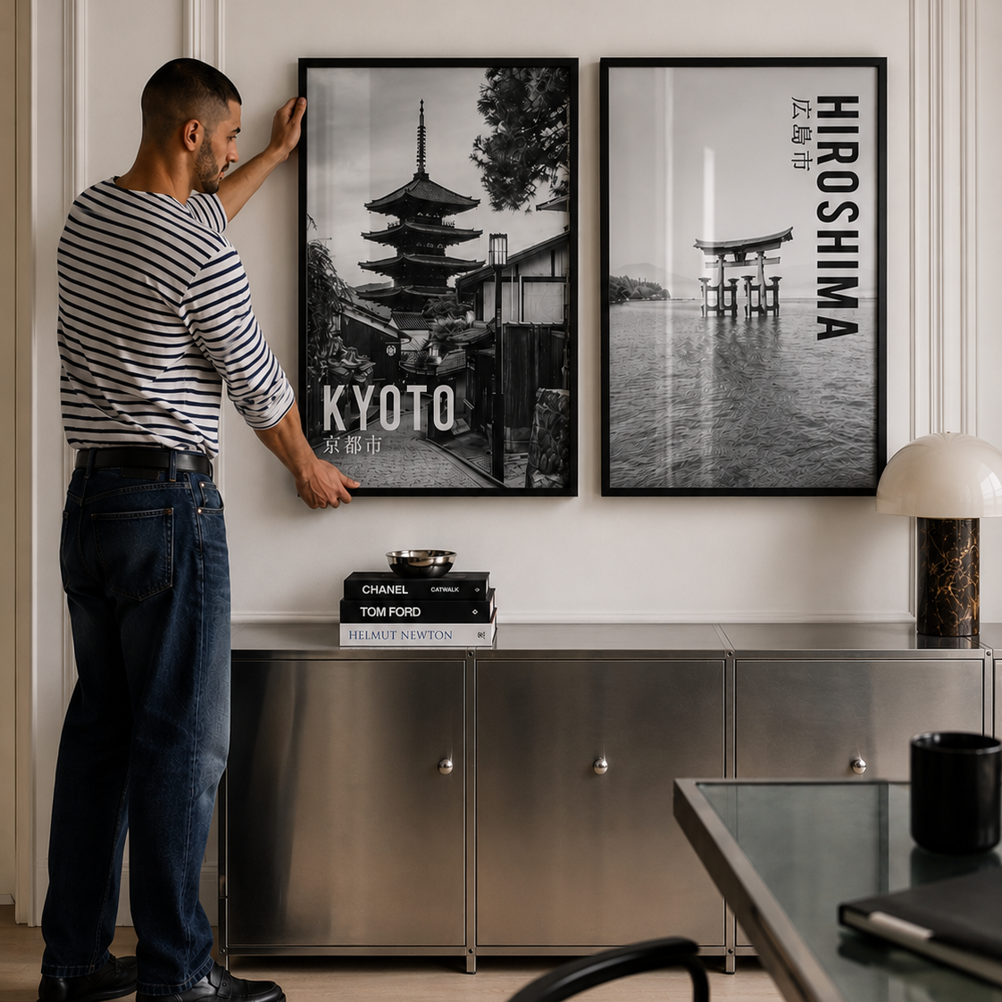 A man in a striped shirt and jeans is adjusting a black-framed wall art print depicting a Kyoto pagoda, next to another framed print with the text 'HIROSHIMA'.