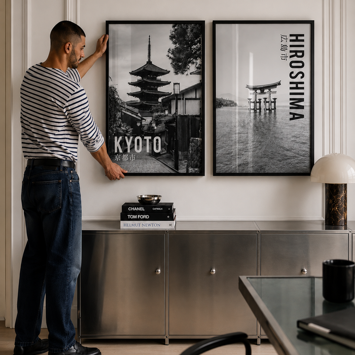 A man in a striped shirt and jeans is adjusting a black-framed wall art print depicting a Kyoto pagoda, next to another framed print with the text 'HIROSHIMA'.