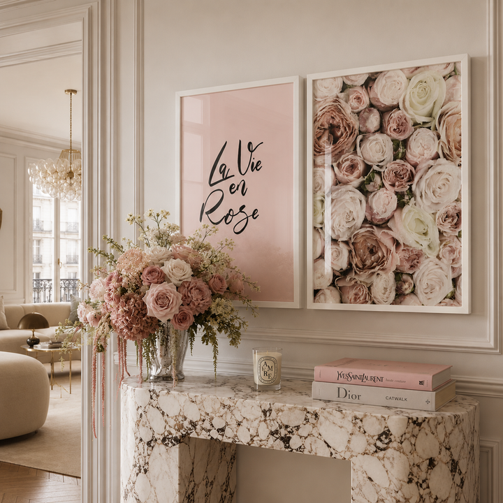 Two framed wall art prints displayed above a marble console table in a styled interior. The left print has a soft blush pink background with black script reading “Le Vie en Rose,” and the right print shows a dense arrangement of pink, blush, cream, and taupe roses.