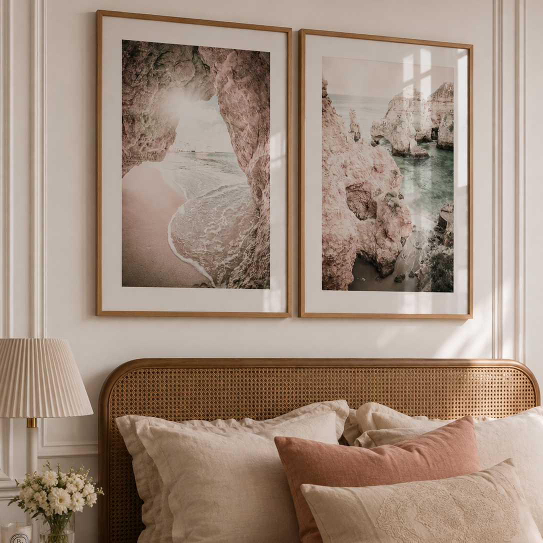 Two framed landscape art prints with pink and beige tones hang above a bed with beige and dusty rose pillows in a softly lit bedroom.