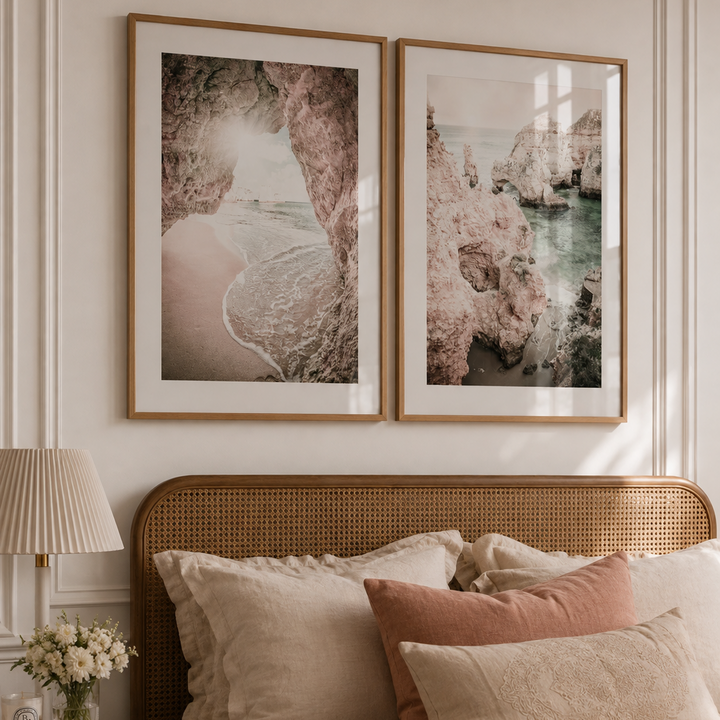 Two framed landscape art prints with pink and beige tones hang above a bed with beige and dusty rose pillows in a softly lit bedroom.