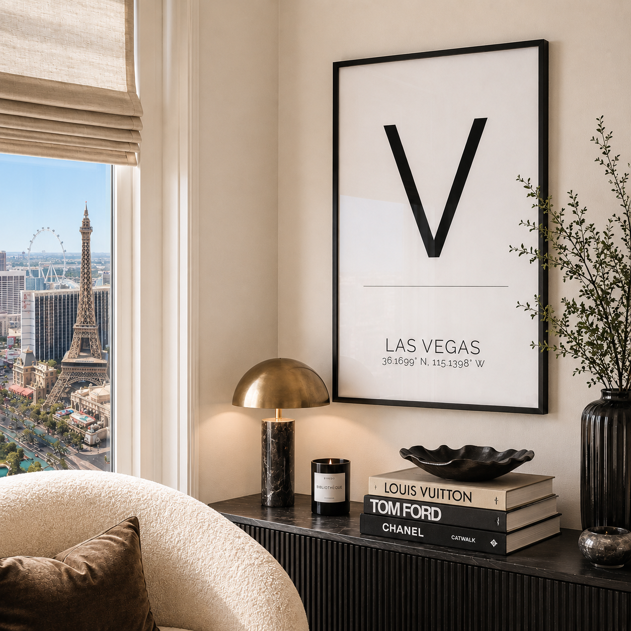 A framed minimalist Las Vegas art print with a large black letter V hangs on a beige wall above a console table. The room includes a window with a view of the Eiffel Tower and Las Vegas skyline, a gold dome lamp, stacked books, and a vase with branches.