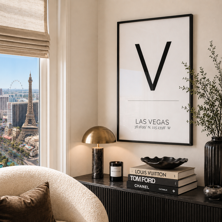 A framed minimalist Las Vegas art print with a large black letter V hangs on a beige wall above a console table. The room includes a window with a view of the Eiffel Tower and Las Vegas skyline, a gold dome lamp, stacked books, and a vase with branches.