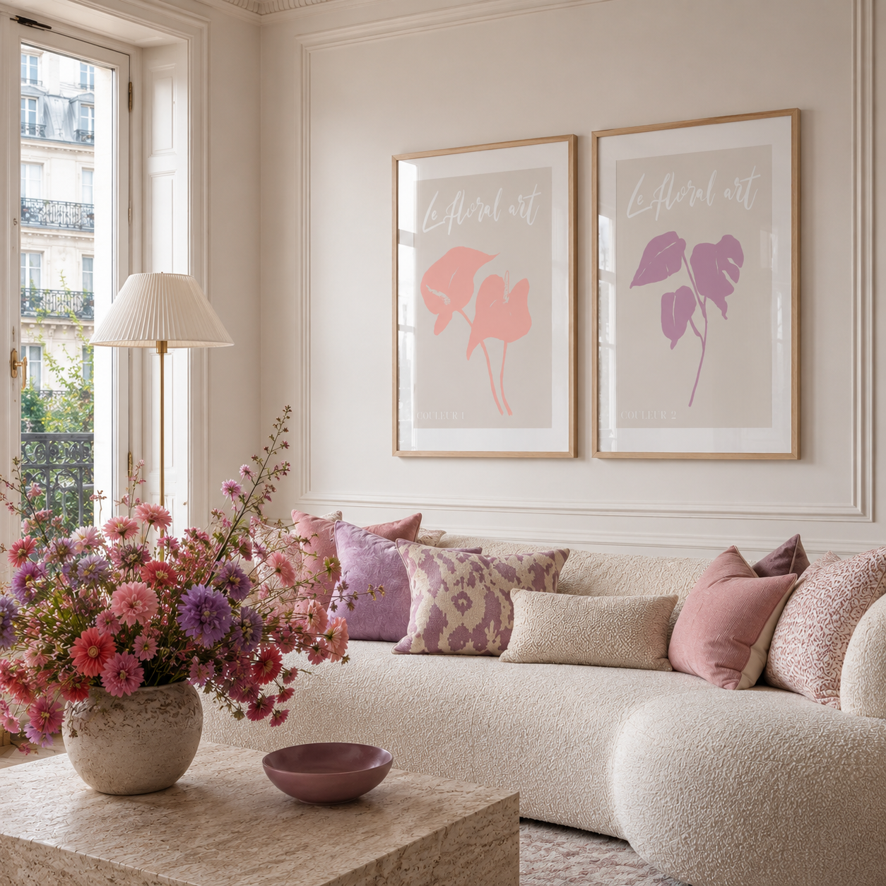 A styled living room scene with a cream boucle sofa, pink and lavender pillows, a floral arrangement on a stone table, and two framed botanical art prints on the wall above the sofa.