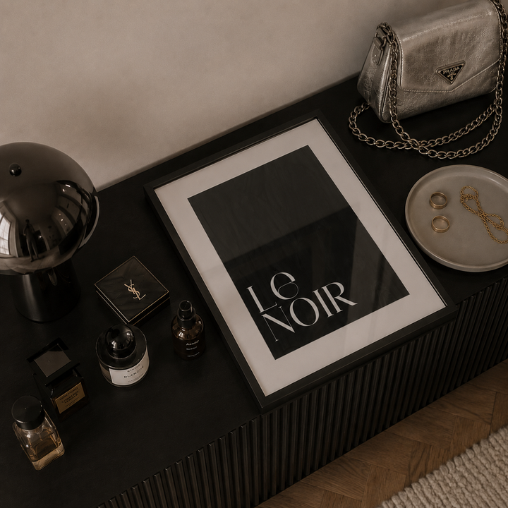 A framed black and white art print with the text 'LE NOIR' in a bold serif font, styled on a dark ribbed console table alongside fashion accessories and perfume bottles.