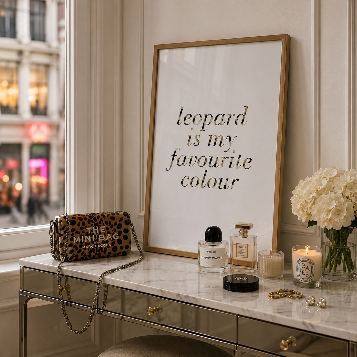 A framed typographic wall print reading 'leopard is my favourite colour' stands on a marble vanity table beside a leopard-print mini bag, perfume bottles, a candle, and white flowers. A real interior setting and window are visible, so this is Lifestyle rather than Product/Packaging.