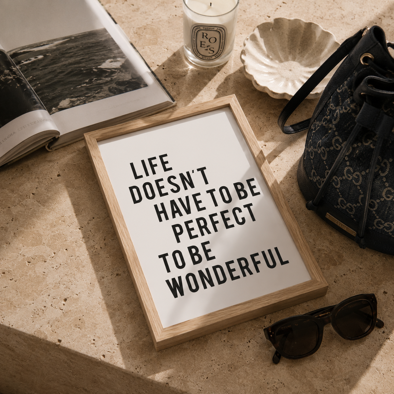 A framed typographic wall art print reading “LIFE DOESN'T HAVE TO BE PERFECT TO BE WONDERFUL” in black text on a white background, styled on a beige stone surface with an open book, candle, shell dish, patterned handbag, and sunglasses nearby.