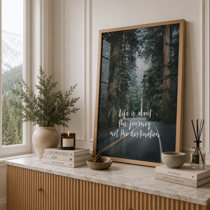 A framed vertical art print leaning on a marble console table in a styled room. The print shows a forest road with the text 'Life is about the journey not the destination' in white script.