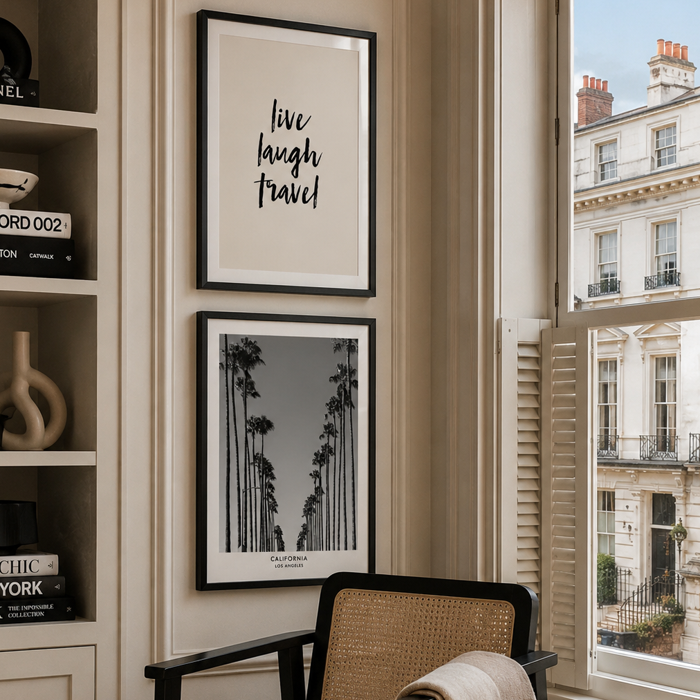 A styled interior corner with two framed wall prints hanging above a black armchair. The upper print reads 'live laugh travel' in black script on a white background, and the lower print is a black-and-white palm tree photograph.