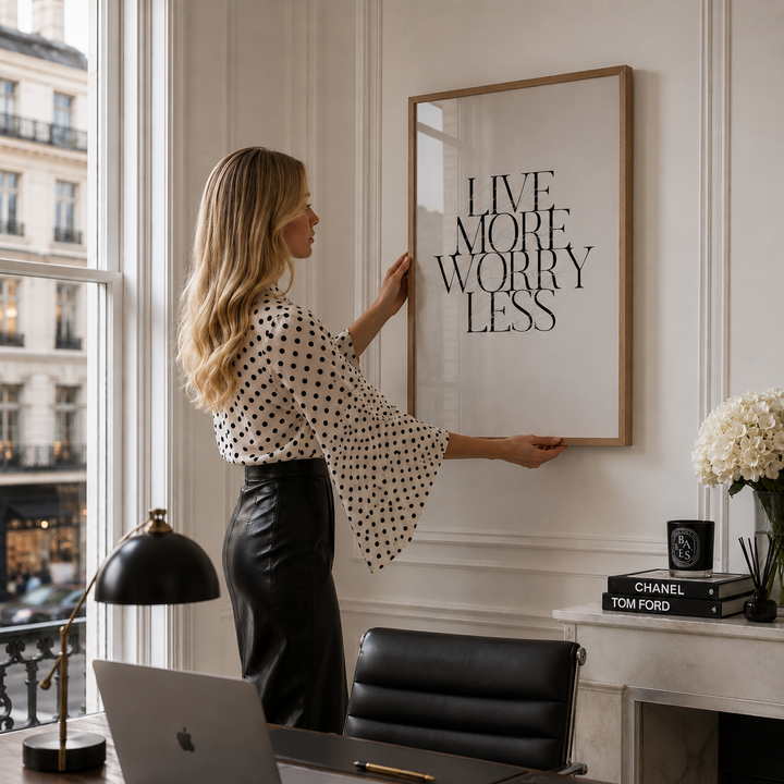 A woman in a black-and-white polka dot blouse and black skirt hangs a large framed typographic wall print reading “LIVE MORE WORRY LESS” in a bright office or home interior. The scene includes a desk, laptop, chair, lamp, books, and flowers.