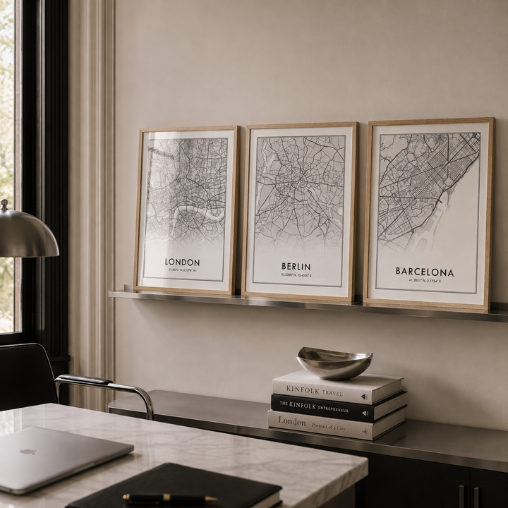 Three framed minimalist city maps of London, Berlin, and Barcelona hang on a beige wall above a shelf in a modern home office setting.