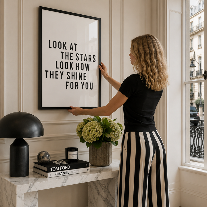 A woman in a black top and black-and-white striped skirt hangs a large black-framed wall art print with the text 'LOOK AT THE STARS LOOK HOW THEY SHINE FOR YOU' on a cream paneled wall in a bright room.