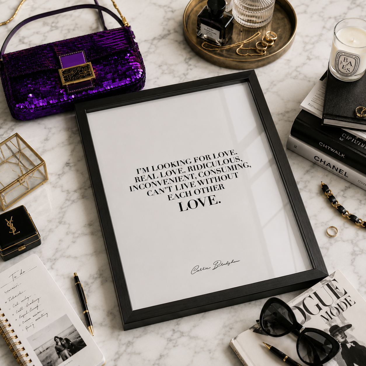 A framed typographic art print with a black quote on a white background, styled in a flat lay on a marble surface with a purple sequined handbag, books, sunglasses, a candle, and other accessories around it.