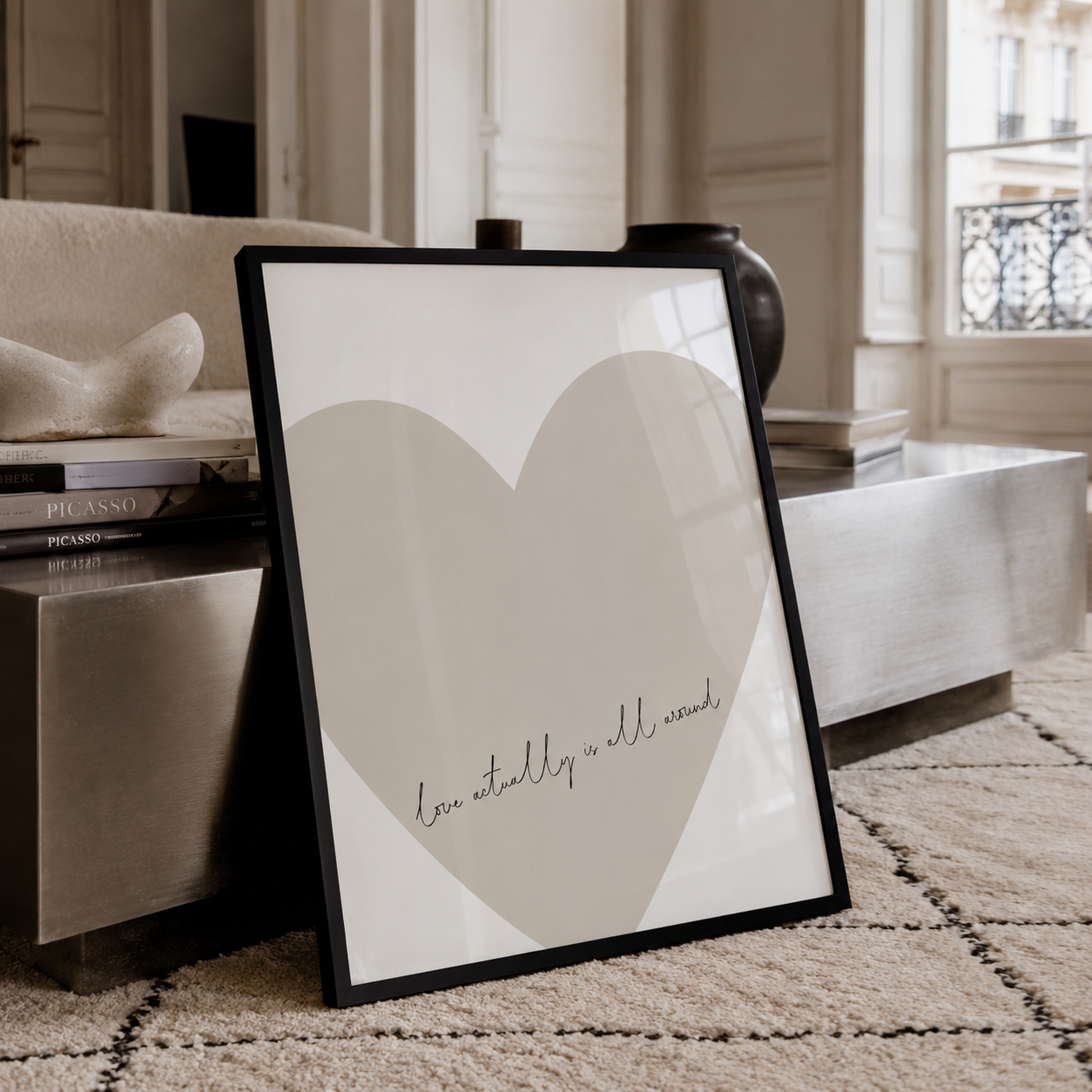 A framed minimalist art print leaning on a beige rug in a modern living room setting. The print features a large light gray heart on a white background with the cursive text 'love actually is all around' at the bottom.