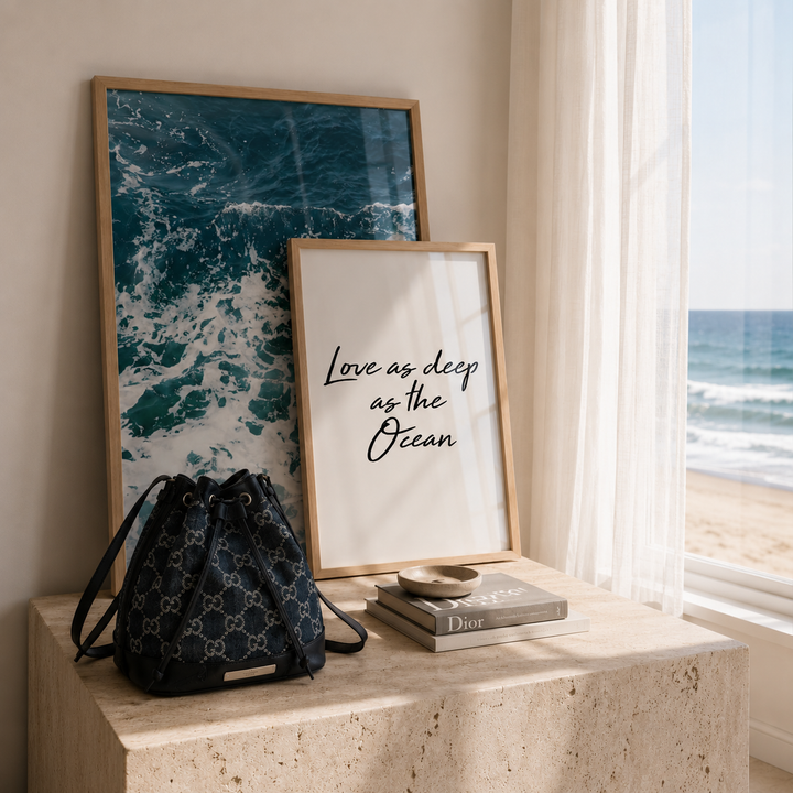 A styled interior scene by a window showing two framed art prints and a black patterned drawstring bag on a stone side table, with an ocean view outside.
