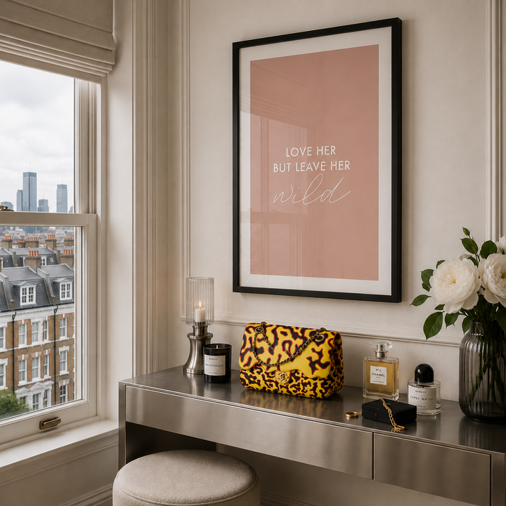 A framed art print with the text 'LOVE HER BUT LEAVE HER wild' in white on a dusty pink background, hanging on a cream wall above a vanity table decorated with a yellow leopard print pouch, perfume bottles, and white roses.