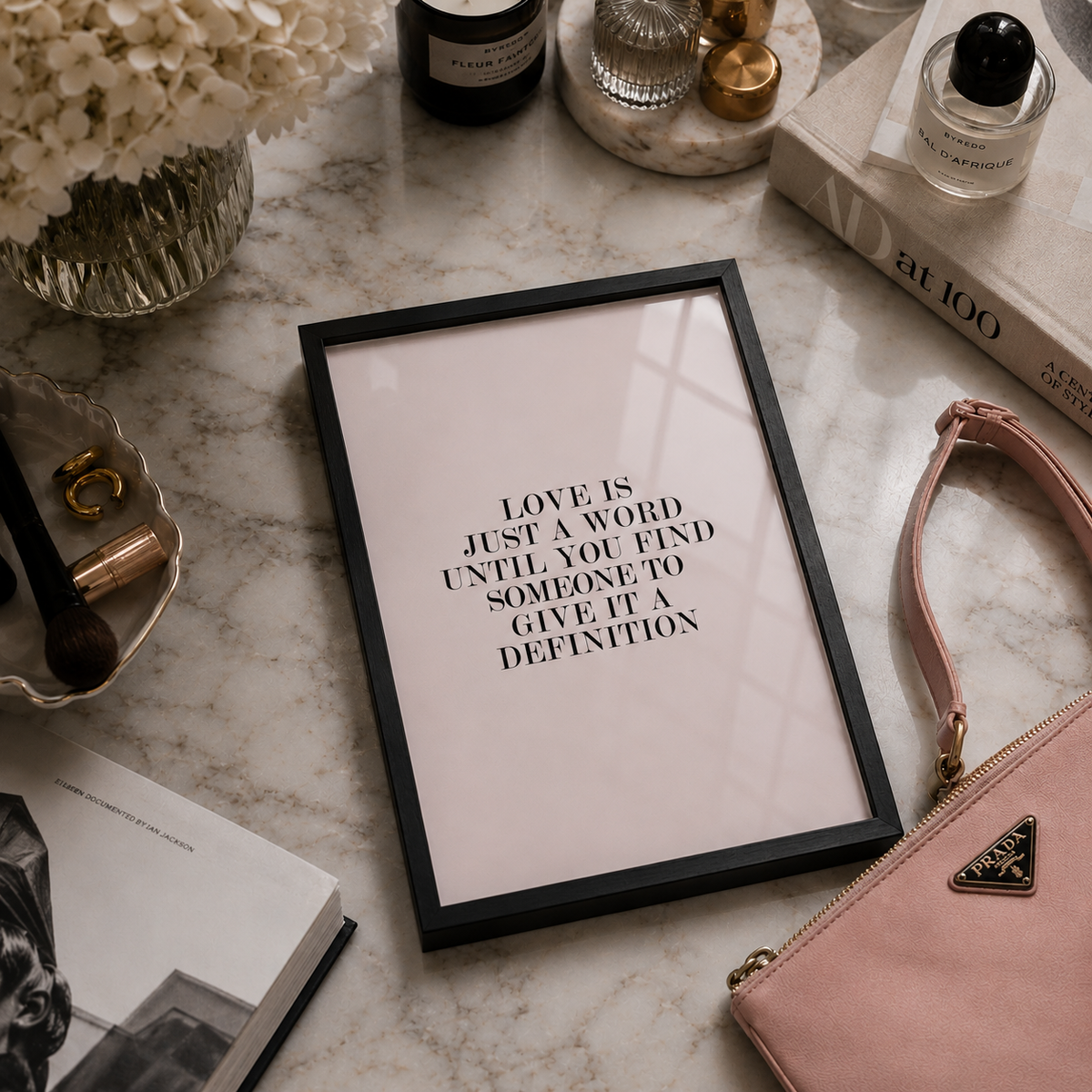 A framed typographic wall art print with the quote 'LOVE IS JUST A WORD UNTIL YOU FIND SOMEONE TO GIVE IT A DEFINITION' in black text on a pale blush background, styled on a marble surface with books, perfume bottles, flowers, and a pink clutch nearby.