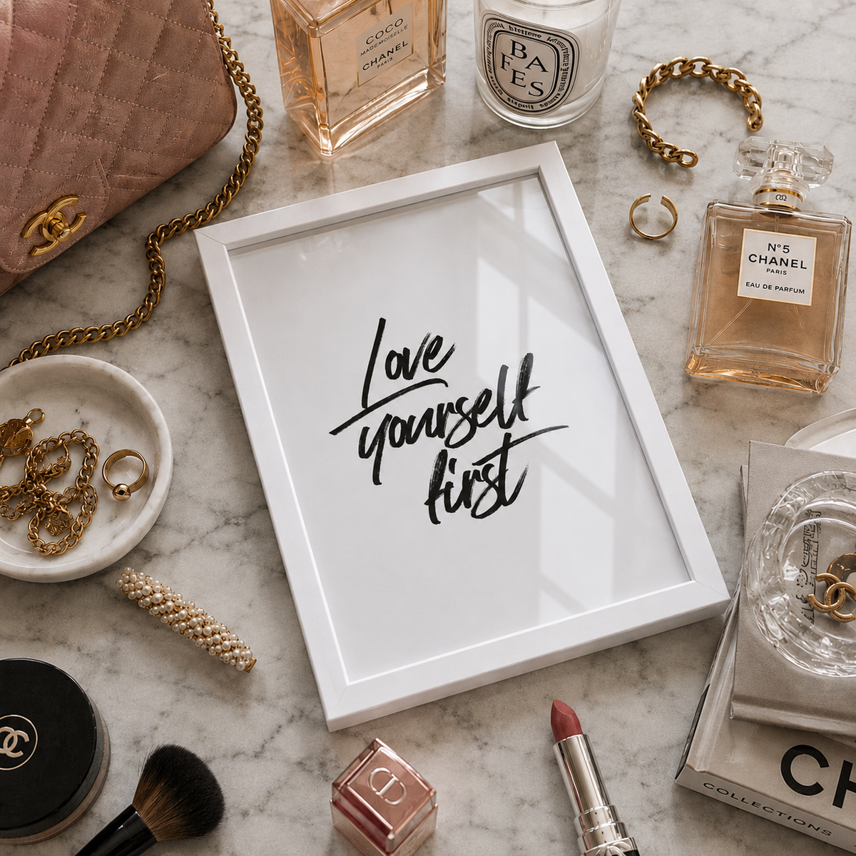 A white framed art print with the phrase 'Love yourself first' in black script, styled in a flat lay on a marble surface surrounded by Chanel products and accessories.