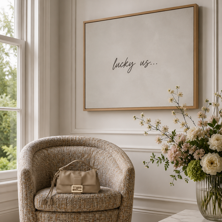 A styled interior scene with the Eichholtz Catene Swivel Chair in Mademoiselle Beige beside a window, a matching beige handbag resting on the chair, and a large luxury oak framed canvas wall print reading 'lucky us.