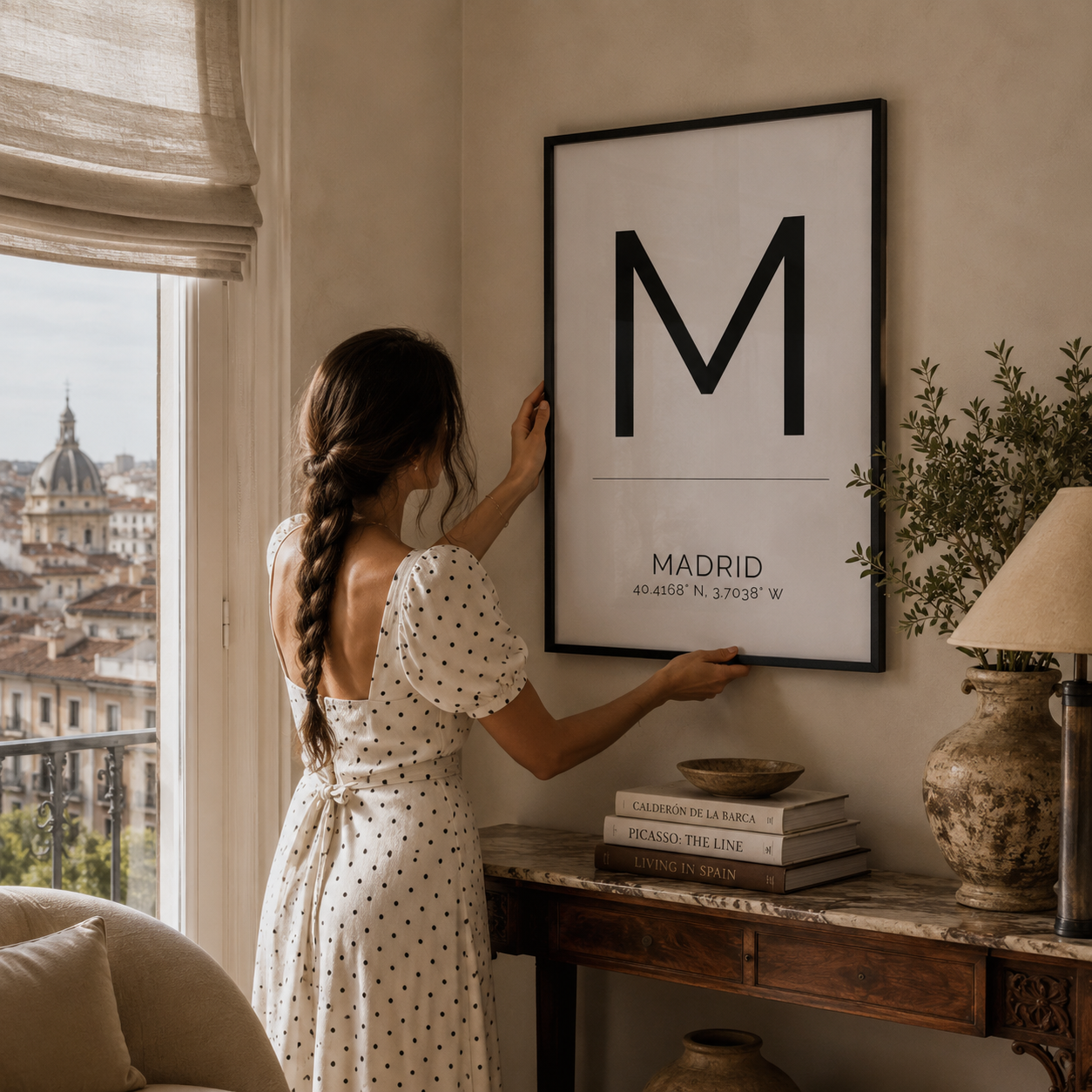 A woman in a white polka-dot dress hangs a large black-framed minimalist poster on a beige wall in a styled room. The poster features a large black letter 'M' with 'MADRID' and coordinates printed below.