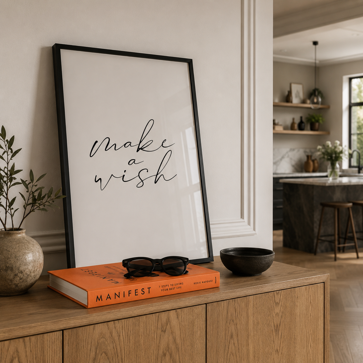 A framed wall art print with the words 'make a wish' in black script on a white background, leaning on a wooden cabinet in a styled home interior. An orange MANIFEST book, sunglasses, and a black bowl sit in front of the frame.