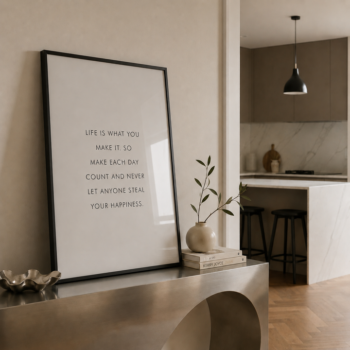 A framed typographic wall art print leaning on a metallic console table in a modern interior. The print has a black frame and black text reading, 'LIFE IS WHAT YOU MAKE IT, SO MAKE EACH DAY COUNT AND NEVER LET ANYONE STEAL YOUR HAPPINESS.