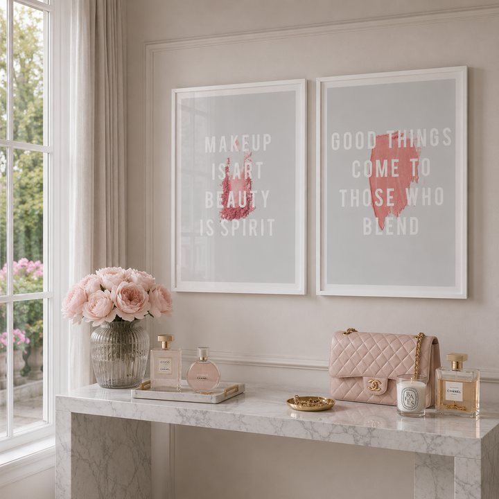 Two framed wall art prints with white text and pink abstract brushstrokes hang above a marble console table in a softly lit room. The table holds pink flowers, perfume bottles, a quilted pink handbag, and small decorative items.