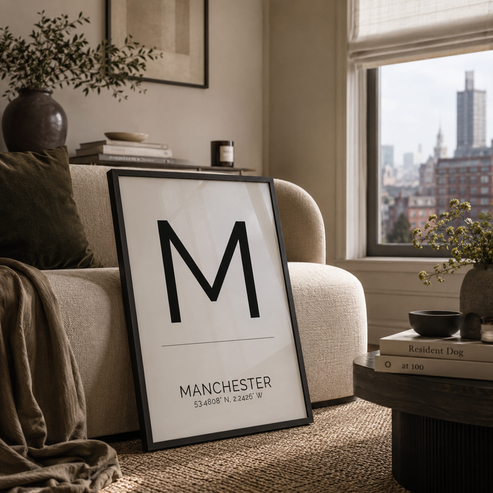 A framed minimalist art print leaning against a beige sofa in a modern living room. The print features a large black letter 'M' with 'MANCHESTER' and coordinates printed below on a white background.