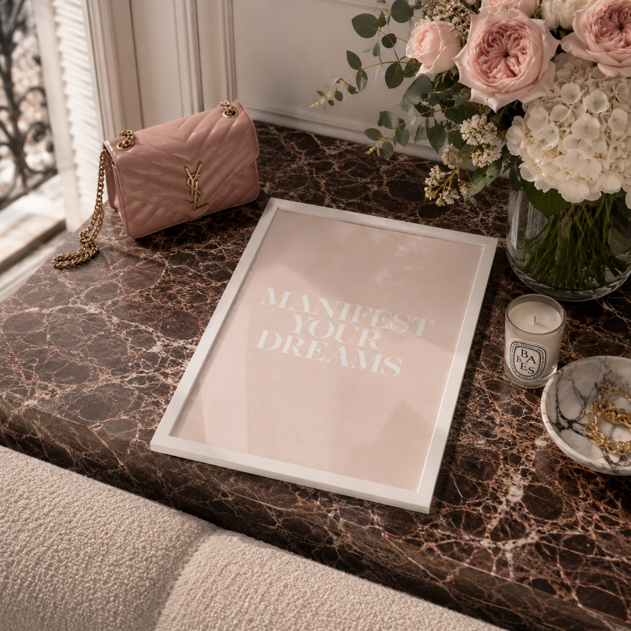 A framed art print with the text 'MANIFEST YOUR DREAMS' in white on a pale blush pink background, styled on a dark marble table next to a pink quilted handbag, a flower arrangement, a candle, and a small dish.