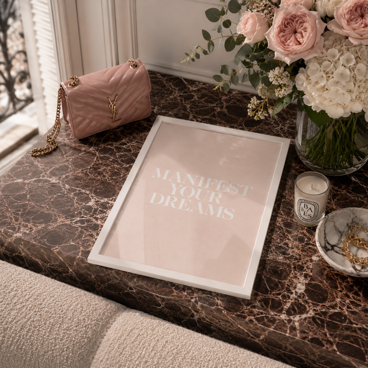 A framed art print with the text 'MANIFEST YOUR DREAMS' in white on a pale blush pink background, styled on a dark marble table next to a pink quilted handbag, a flower arrangement, a candle, and a small dish.