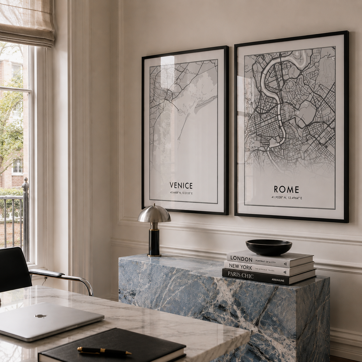 Two framed black-and-white city maps, Venice and Rome, displayed side by side on a cream wall in a styled home office setting with a blue marble console table and a window.