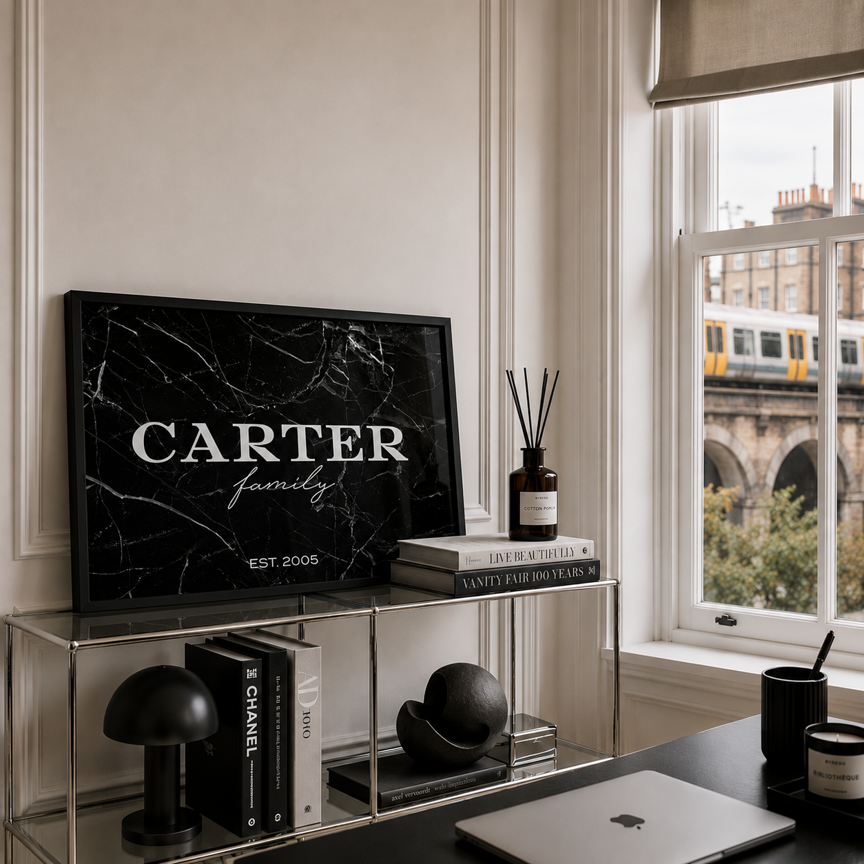 A personalised framed black marble-style wall art print with the text 'CARTER family EST. 2005' displayed on a glass shelf in a modern interior beside a window with an outdoor view.