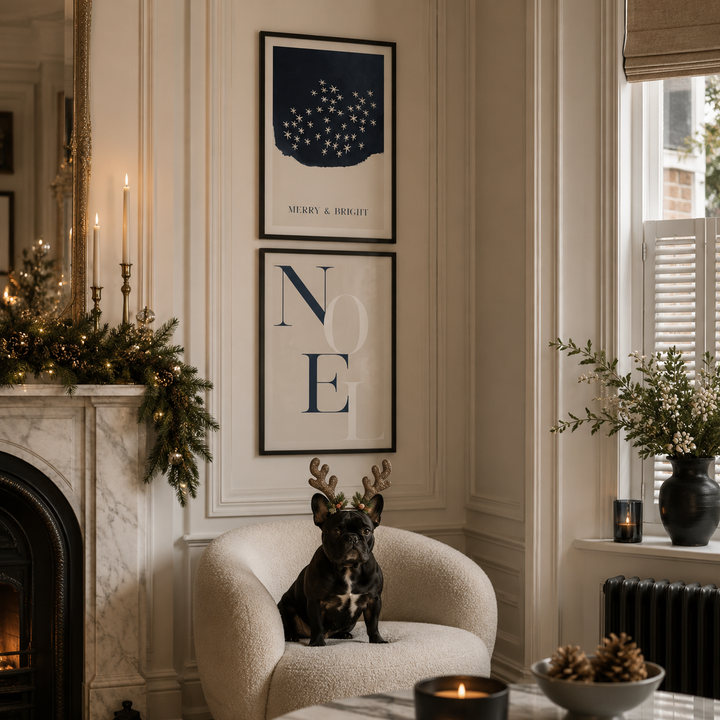 A black French bulldog wearing small reindeer antlers sits on a cream armchair in a decorated living room. Two framed wall art prints hang above the chair, one with a navy blue abstract shape and stars and the text 'MERRY & BRIGHT', and the other with large navy letters spelling 'NOEL' on a beige background.