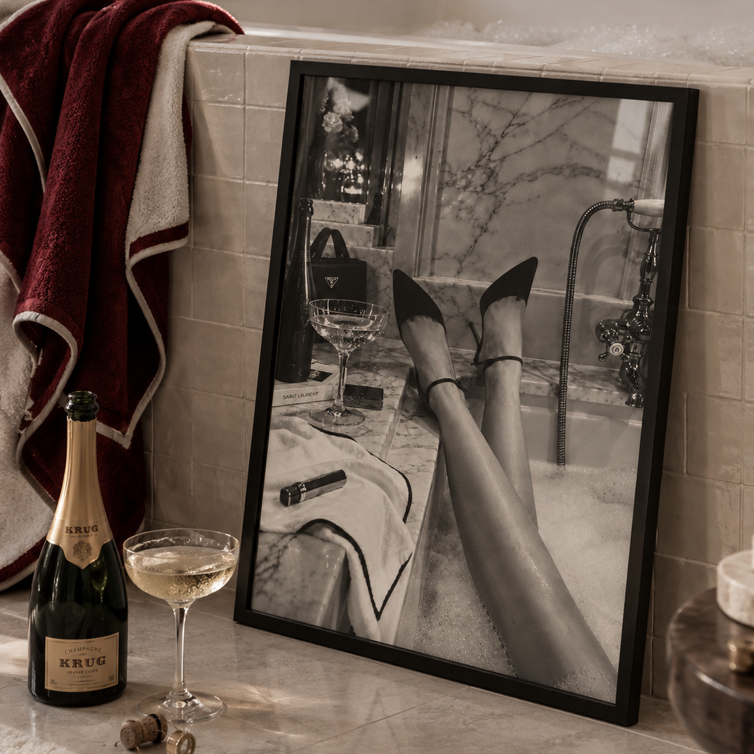 Black pointed-toe high heels with ankle straps worn by a person reclining in a bubble bath, with marble walls and a cocktail glass nearby.