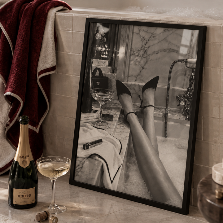 Black pointed-toe high heels with ankle straps worn by a person reclining in a bubble bath, with marble walls and a cocktail glass nearby.