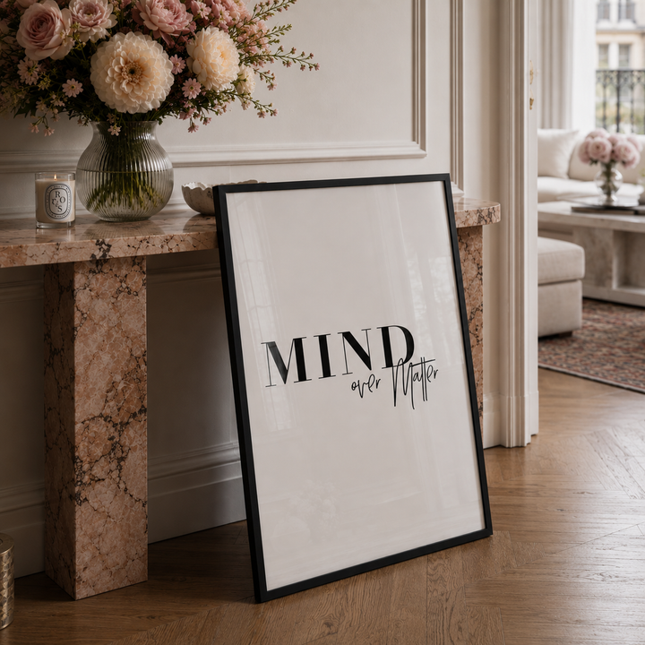 A framed typographic wall art print leaning against a marble console table in a bright living room. The print has a white background with the words “MIND over Matter” in black serif and script lettering, and a thin black frame.