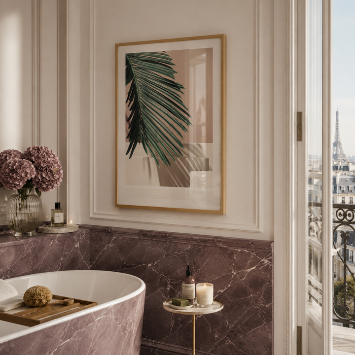 A framed botanical art print featuring a green palm leaf on a beige and white background, styled in a gold frame on a bathroom wall above a marble tub and side table.