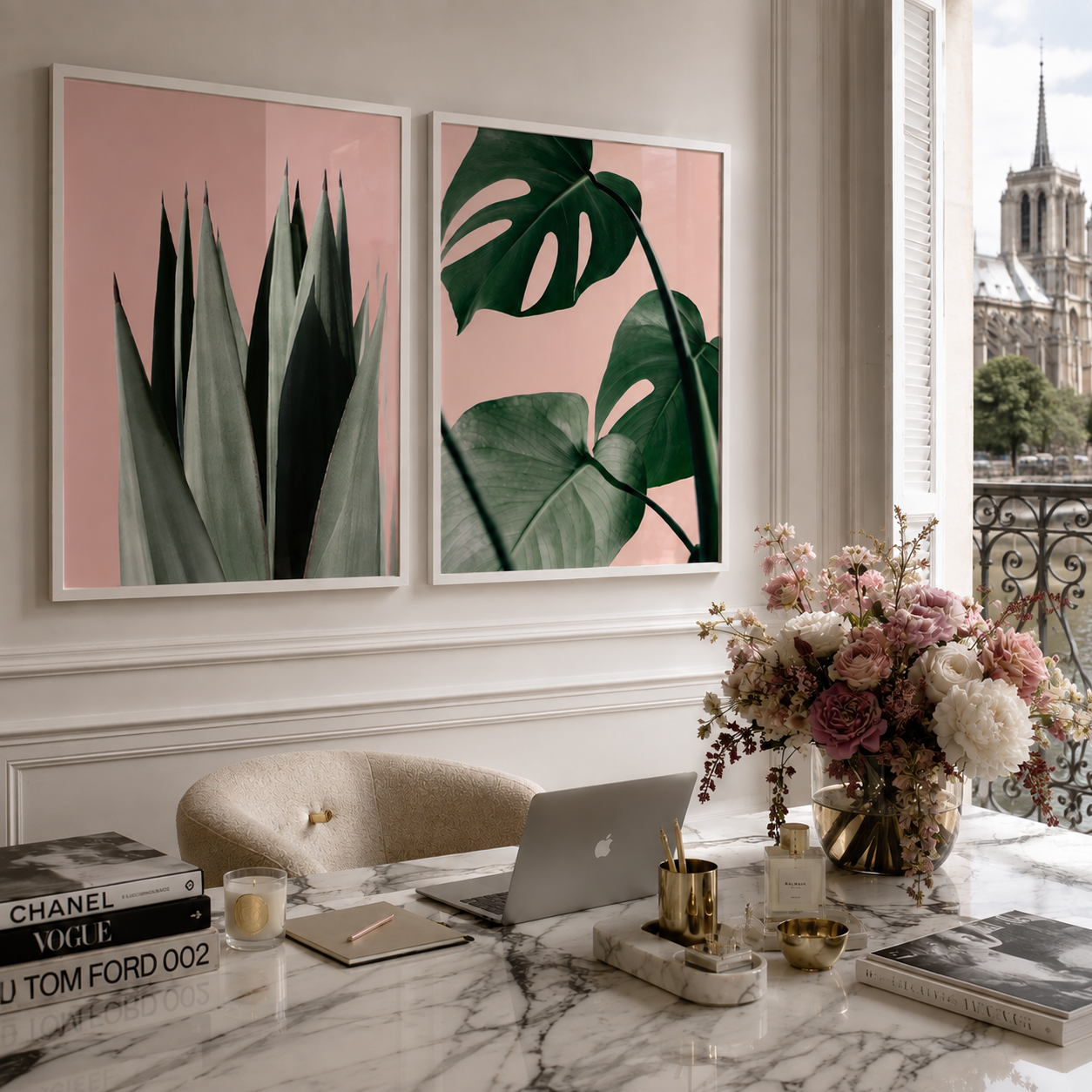 Two framed botanical art prints hang on a light beige wall above a marble desk in a styled home office. The left print shows dark green succulent leaves on a dusty pink background, and the right print shows large green monstera leaves on the same pink background.