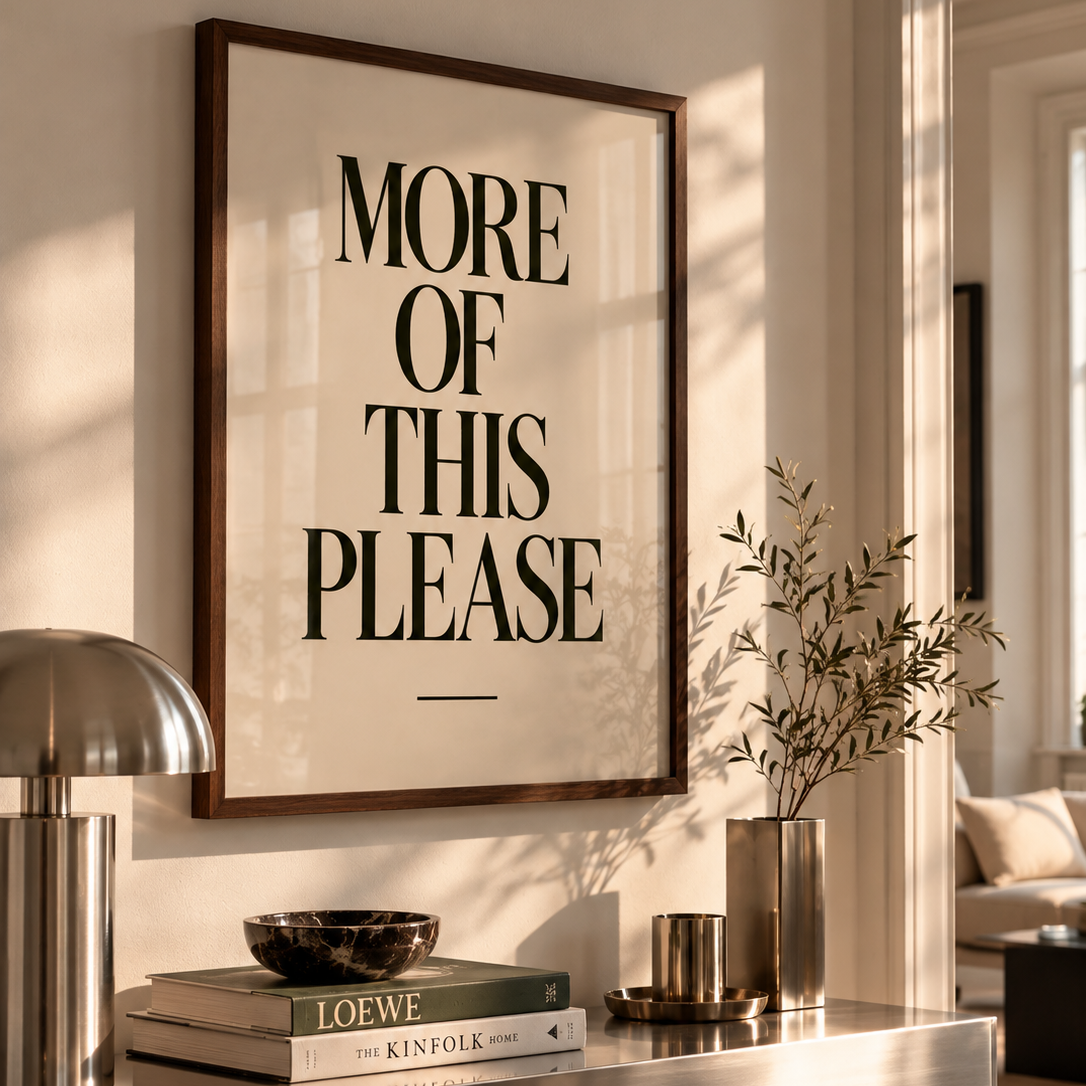 A framed typographic wall art print reading “MORE OF THIS PLEASE” hangs on a beige wall above a console table. The dark wood frame is styled in a warm, modern interior with a silver lamp, books, a bowl, and a vase with branches.