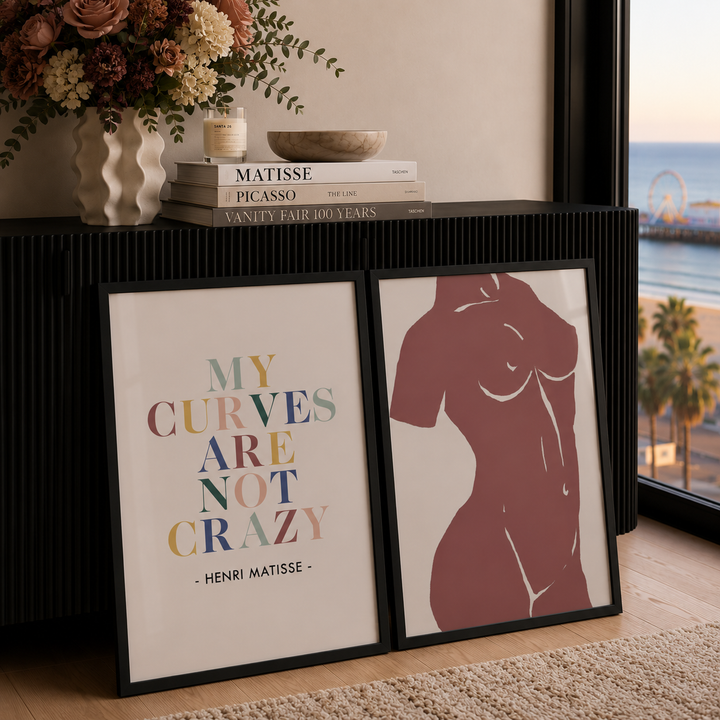 Two framed art prints leaning against a black ribbed console table in a styled interior. The left print features a Henri Matisse quote in multicolored lettering, and the right print shows a minimalist maroon female torso silhouette.