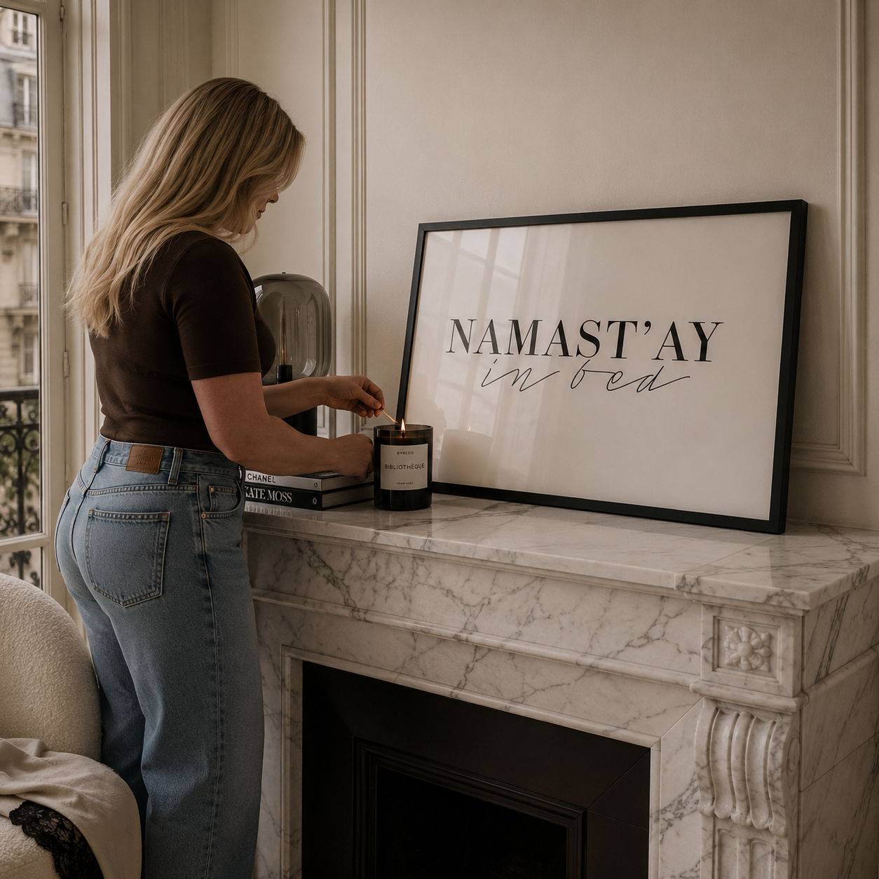 Person arranging books and a candle on a marble fireplace with a framed sign reading 'Namast'ay in bed'.